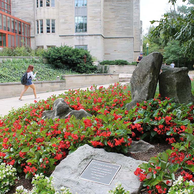 A student walks by Jordan Hall. Red begonias bloom in a flowerbed in the foreground.