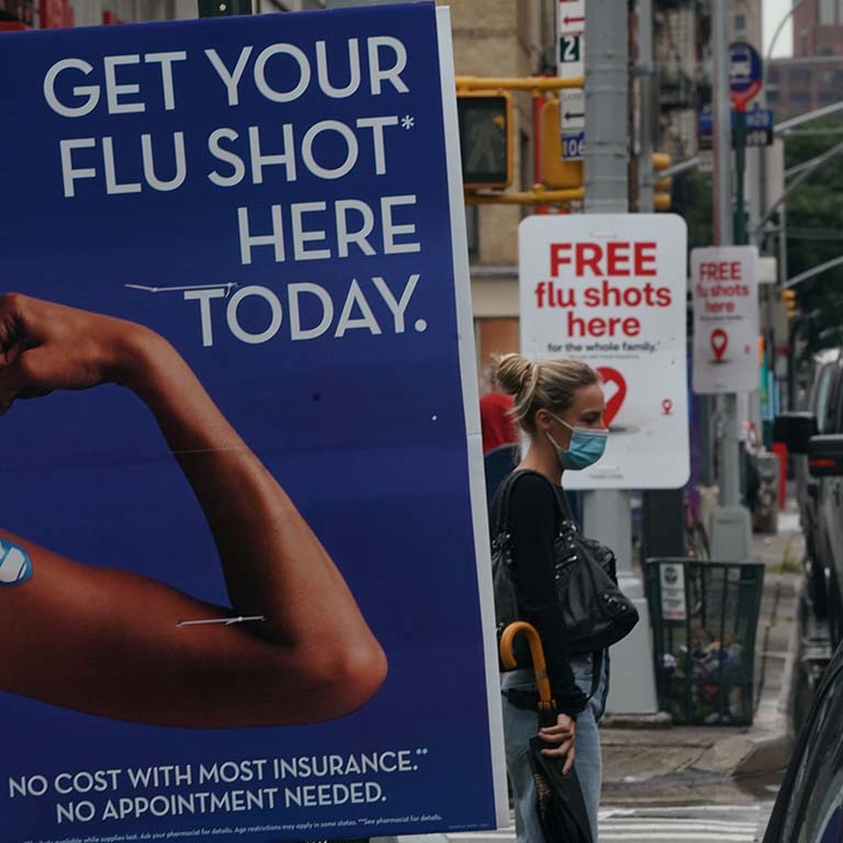 Signs encouraging people to get their flu shot are displayed on light posts along a street as a woman wearing a face mask waits to cross the traffic.