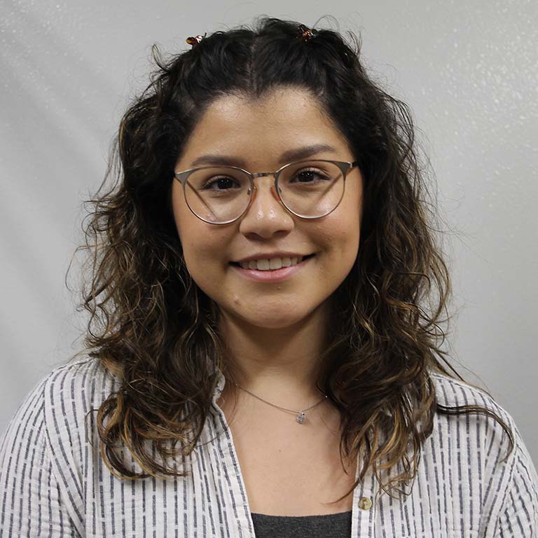 Victoria Lydick--a white woman with long, wavy, dark brown hair and wearing glasses--smiles in the portrait photo.