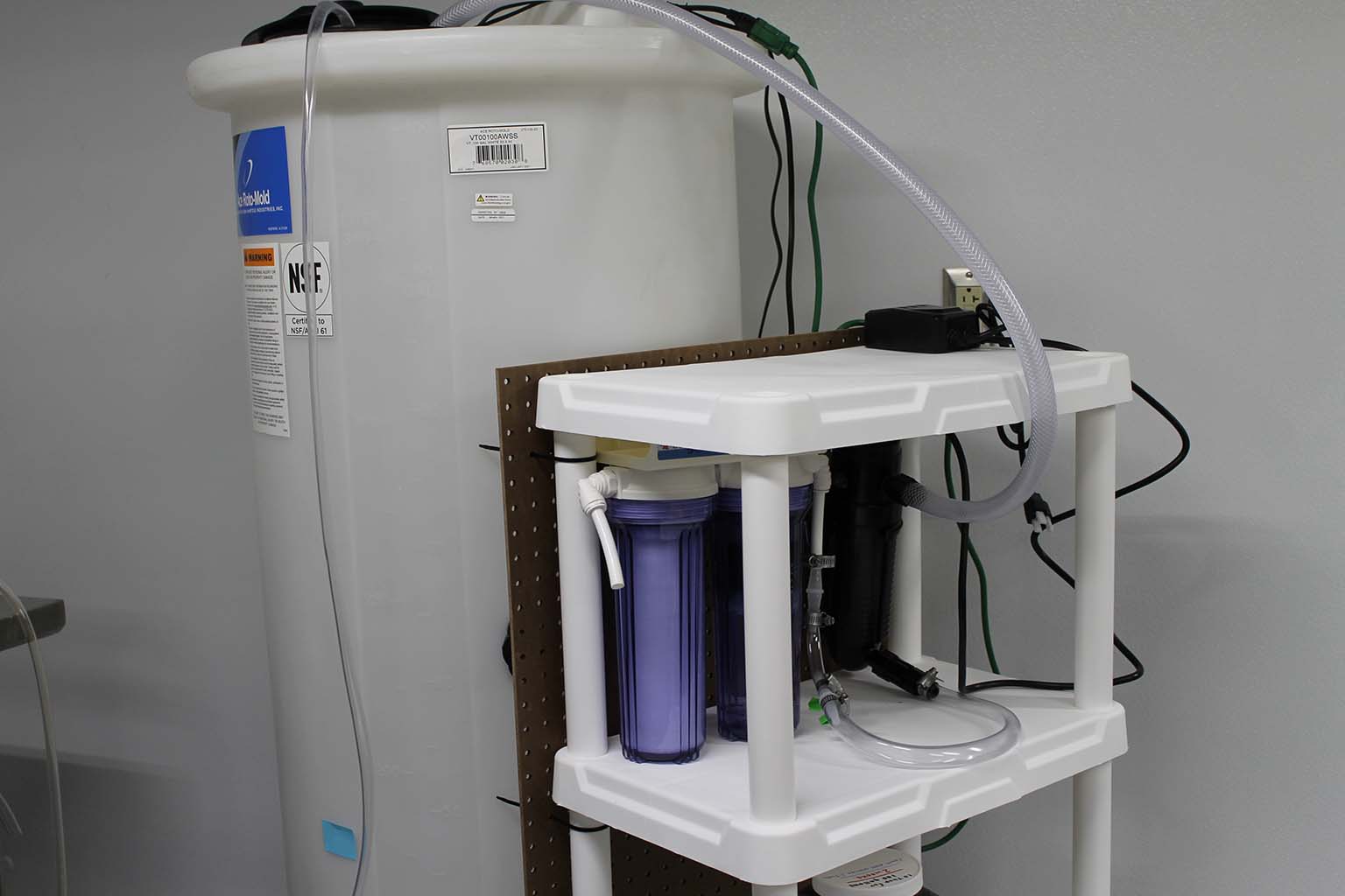 A tall, cylindrical, white, 100-gallon tank that holds the artificial seawater for the coral tanks is connected to three filter containers on a shelf next to the tank. 