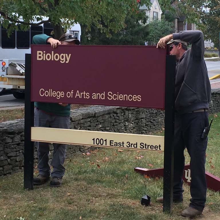 Two men slide a large red sign into place between two vertical posts. The sign reads: Biology, College of Arts and Sciences, 1001 East 3rd Street.  The sign is being installed in the lawn in front of the newly named Biology Building as traffic passes by along 3rd Street.